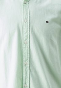 Camisa de botones verde claro hecha de una tela suave, con una sutil textura y un pequeño logo bordado en el pecho, botones blancos.