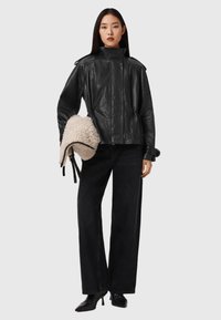 Zwarte leren jas met hoge kraag, vooraanrits en uitgesproken schoudernaden. Model houdt een crèmekleurige neplederen handtas vast. Zwarte broek met wijde pijpen.