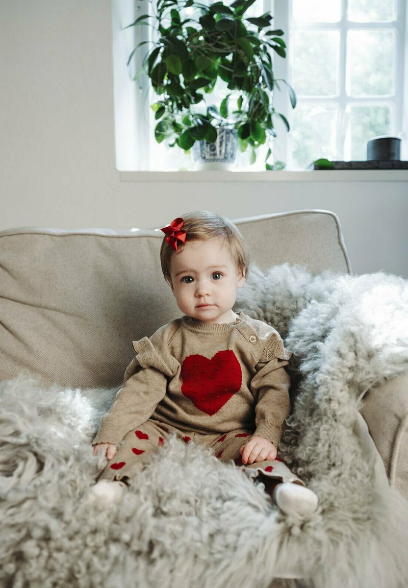 Maglione beige a maglia con un grande cuore rosso, abbinato a pantaloni rossi e beige con fantasia. Bambino seduto su una coperta morbida grigia chiara.