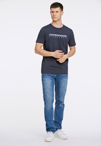 T-paita tummansinisellä värillä, jossa on valkoinen "COPENHAGEN" -teksti, lyhyet hihat, yhdistetty vaaleansinisiin farkkuihin ja valkoisiin lenkkareihin, seisoo yksinkertaisella taustalla.