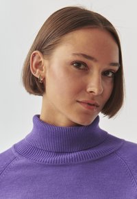Pull-over en col roulé violet avec une texture côtelée et des détails de couture. Le mannequin a les cheveux courts et porte des boucles d'oreilles en or.