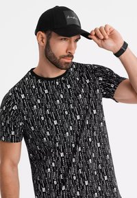 Camiseta negra con patrón de texto blanco vertical; mangas cortas, escote redondo. Gorra negra con un parche de logo mate, llevada de lado.