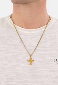 Collana in oro con un pendente a croce testurizzato e con motivi intagliati, appoggiata su una camicia bianca semplice.