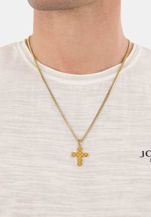 Collier en chaîne dorée avec un pendant croix texturé présentant des motifs découpés, reposant sur une chemise blanche unie.