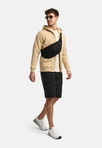 Haut à capuche crème zippé, sac bandoulière noir, chemise légère, shorts noirs, baskets blanches et grises, lunettes de soleil. Style streetwear décontracté.