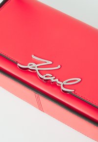 Clutch in pelle rossa con dettaglio del logo argentato. Presenta una texture liscia, bordi neri a contrasto e una forma rettangolare.