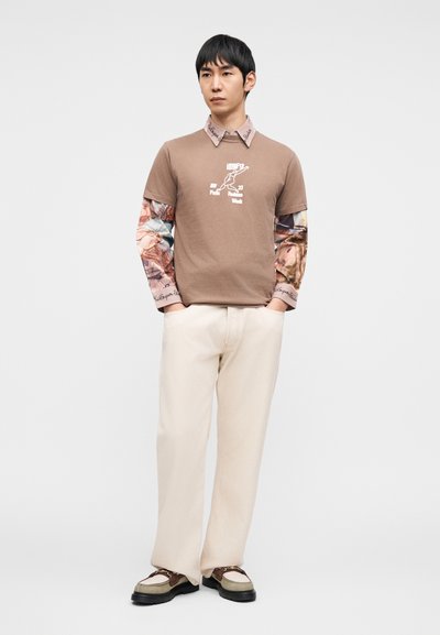 Καφέ κοντομάνικο πουκάμισο με γραφικό σχέδιο, φορεμένο πάνω από ένα μακρυμάνικο πουκάμισο με λουλουδάτο μοτίβο. Παντελόνια σε εκρού χρώμα και πολύχρωμα παπούτσια.