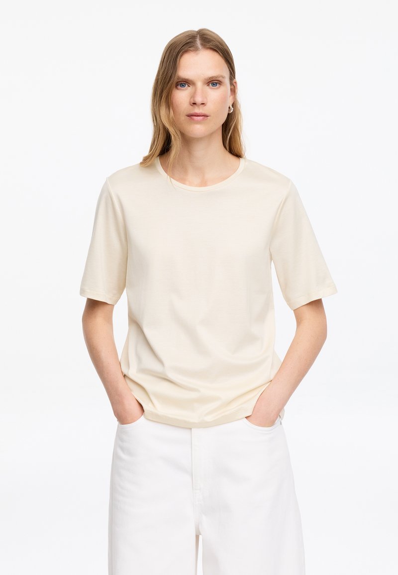 Femme aux cheveux blonds et raides, portant une chemise crème à manches courtes et un pantalon blanc, debout les mains dans les poches devant un fond uni.