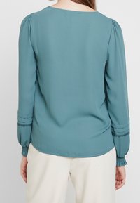 Blouse bleu sarcelle à manches longues, col rond et détails texturés aux poignets ; tissu lisse avec une légère brillance et plis doux aux épaules.