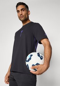 Zwarte t-shirt met korte mouwen en paarse accenten, ronde halslijn, lichtgewicht stof. Model houdt een witte en blauwe voetbal vast.
