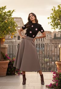 Czarny top z krótkim rękawem z różowym haftem kwiatowym, w połączeniu z beżową spódnicą o wysokim stanie i brązowymi botkami za kostkę; plener z roślinami.