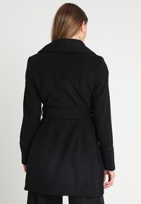 Cappotto nero con colletto, vita infasciata e maniche lunghe. Realizzato in un tessuto testurizzato con linee pulite e senza hardware visibile.