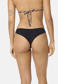 Mujer con braguitas de bikini negras y un top de bikini estampado atado por detrás, mostrando un tatuaje en su brazo izquierdo.