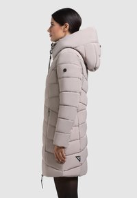 Béžová bundu s kapucňou, má prešívaný dizajn, bočné zipsy a logo nášivku na dolnej prednej časti. Hladký, matný povrch.