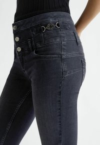 Acercamiento de una persona que lleva puestos unos pantalones ajustados oscuros con tres botones metálicos y un detalle circular en la cintura.