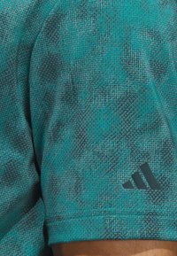 Short-sleeve athletic shirt in teal, featuring a textured pattern and a dark gray Adidas logo on the sleeve. Smooth fabric.