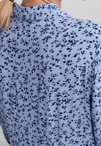 Lyseblå bluse med et blomstermotiv i marineblå, med en blød tekstur og kravedetalje, set bagfra.