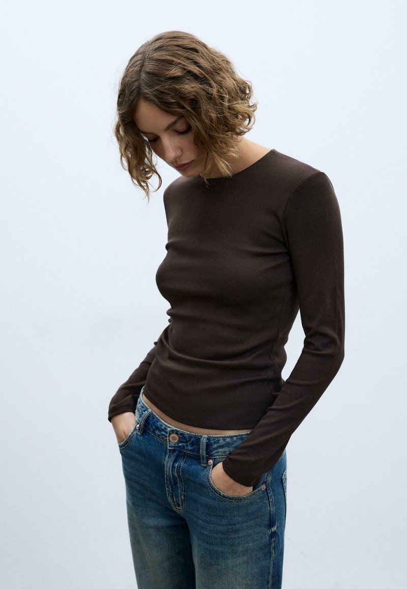 Maglia marrone aderente a maniche lunghe con scollo rotondo, abbinata a jeans blu slavati. Il tessuto appare morbido ed elastico, con una trama liscia.