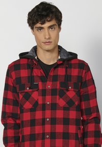 Piros és fekete kockás flannel ing, szürke kapucnival, elülső zsebekkel, gombos záródással és állítható húzózsinórral a kapucnin.