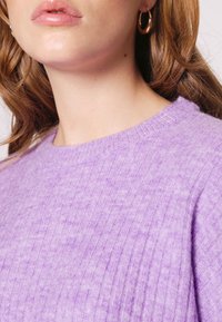 Lila gerippter Pullover mit Rundhalsausschnitt und weichem Material, ergänzt durch kleine goldene Creolen.