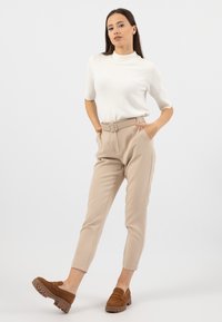 Top in maglia bianco a maniche corte abbinato a pantaloni beige chiaro a sigaretta con vita regolabile. Il modello indossa zoccoli in suede marrone con suole spesse.