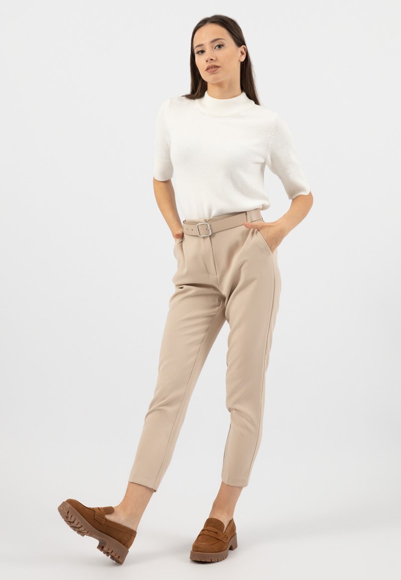 Top in maglia bianco a maniche corte abbinato a pantaloni beige chiaro a sigaretta con vita regolabile. Il modello indossa zoccoli in suede marrone con suole spesse.