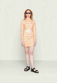 Vestido de mangas longas com padrão laranja e cintura recortada, silhueta ajustada, combinado com sandálias pretas e óculos de sol.