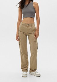 Vrouw in beige cargobroek, witte sneakers en een grijze mouwloze crop top, staande tegen een effen lichte achtergrond.