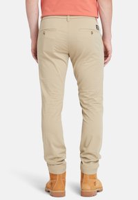 Pantaloni beige slim-fit con tasche laterali e posteriori, dal tessuto liscio. Indossati con stivali alla caviglia marrone chiaro.