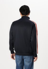 Σακάκι στίβου ναυτικού τύπου με ψηλό γιακά, διαθέτει κόκκινες και λευκές ρίγες στο πλάι, ελαστικούς μανσέτες και στρίφωμα; λεία υφή. Φορεμένο με μπλε τζιν.