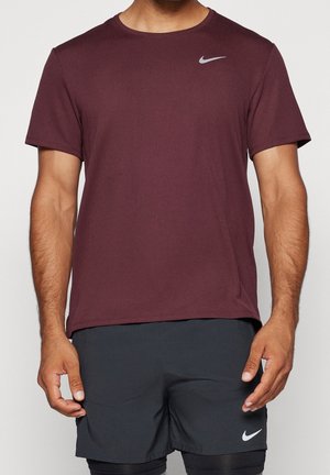 Homme portant un t-shirt Nike bordeaux à manches courtes et un short Nike noir, debout devant un fond clair uni.