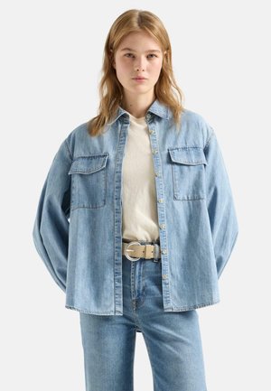 Young woman wearing light blue denim shirt and jeans with beige belt and light beige top, standing with hands behind back.