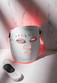 Witte LED-gezichtsmasker met label STYLPRO en roodlichtgloed, verbonden met een witte afstandsbediening op een marmeren oppervlak.