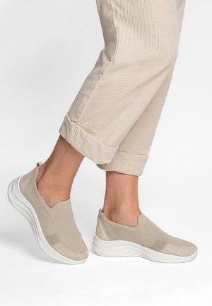 Legs wearing beige corduroy rolled-up pants and slip-on beige knit sneakers with white thick soles on a plain white background.