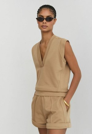 Femme portant un haut sans manche couleur beige avec un col en V, un short assorti avec un ourlet roulé, des lunettes de soleil noires en forme d’œil de chat et un bracelet en or, une main dans la poche.