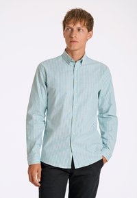 Lichtblauw en wit gestreepte longsleeve shirt met een button-down kraag en knopen aan de voorkant, gemaakt van een textuurstof. Model draagt zwarte broeken.