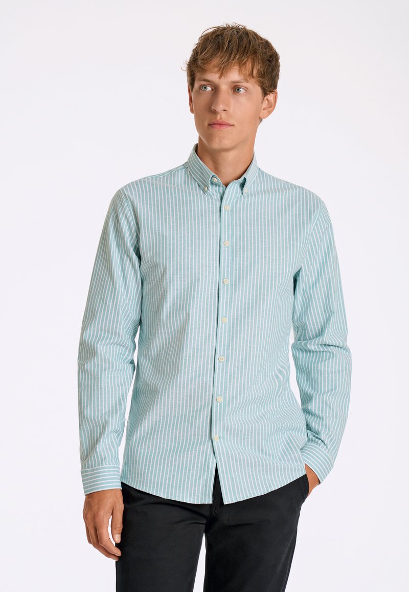 Lichtblauw en wit gestreepte longsleeve shirt met een button-down kraag en knopen aan de voorkant, gemaakt van een textuurstof. Model draagt zwarte broeken.