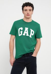Πράσινο βαμβακερό t-shirt με μεγάλο λευκό κείμενο "GAP", κοντά μανίκια και στρογγυλή λαιμόκοψη. Συνδυασμένο με σκούρο μπλε παντελόνι.