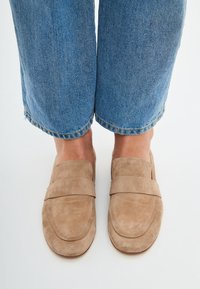 Tan suède loafer heeft een gladde bovenkant met een gestikt ontwerp, een brede band over de wreef en een ronde neus. Draagt met cropped blauwe jeans.