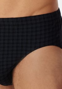 Torse masculin portant un slip noir avec un motif de grille bleu clair, montrant la taille et les zones supérieures de la cuisse.