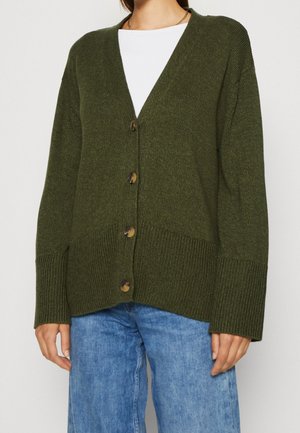 Frau trägt eine olivgrüne, geknöpfte Strickjacke über einem weißen Top mit blauen Jeans, steht vor einem schlichten hellen Hintergrund.