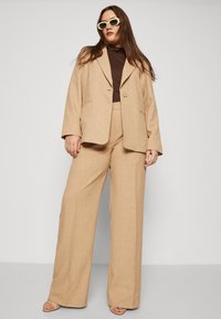 Beige tailored blazer with two buttons, paired with high-waisted wide-leg pants. Brown turtleneck underneath, and green sunglasses.