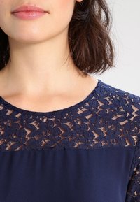 Žena s vlasy po ramena v tmavě modrém topu s květinovou krajkou na horní části hrudníku a ramenou.