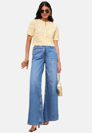 Haut cropped jaune à manches courtes avec des accents texturés, associé à un jean en denim taille haute à jambes larges. Le mannequin tient un sac métallique.