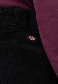 Pantalones de pana negros con finas estrías, que cuentan con un parche del logo de Dickies en el bolsillo trasero. Combinados con una parte superior de color burdeos.