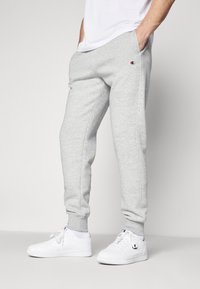 Grijze joggingbroek met zijzakken, elastische tailleband en taps toelopende boorden; logopatch op de linkerheup. Gecombineerd met witte sneakers.