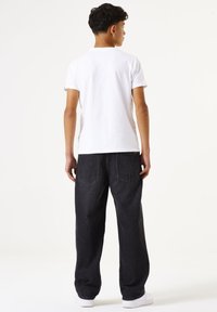 Camiseta de algodón blanca de manga corta, combinada con pantalones vaqueros oscuros y holgados y zapatillas blancas, vista desde atrás.