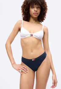 Γυναίκα με σγουρά μαλλιά που φοράει λευκό bralette και σκούρο μπλε εσώρουχα, και τα δύο με ένα μικρό ορθογώνιο λογότυπο στο κέντρο.