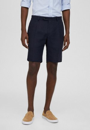 Shorts en lin bleu marine avec des plis sur le devant, une fermeture à bouton et deux poches latérales. Associés à des chaussures slip-on beiges et une chemise à rayures bleu clair.