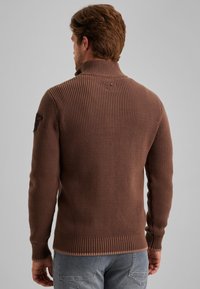 Bruine geribbelde trui met een hoge kraag, voorzien van een textuurgebreide stof en een logo op de linkerschouder. Aansluitende pasvorm met een afgewerkte onderkant.